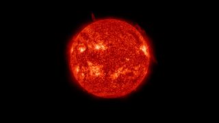 Image of the sun in space. It appears as a dark red/orange sphere with bright areas and a few thin filaments extending into space.